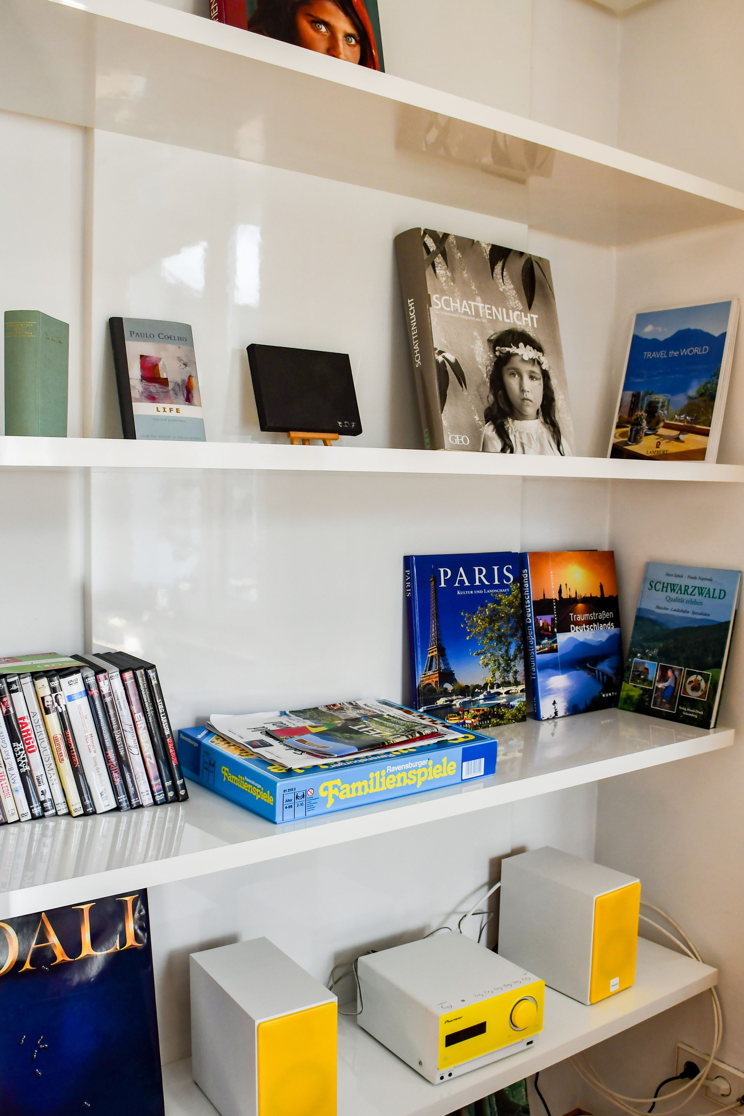 Ein Bücherregal mit verschiedenen Büchern und DVDs, eine kleine Staffelei mit einem Gemälde und zwei Lautsprecherregale.
