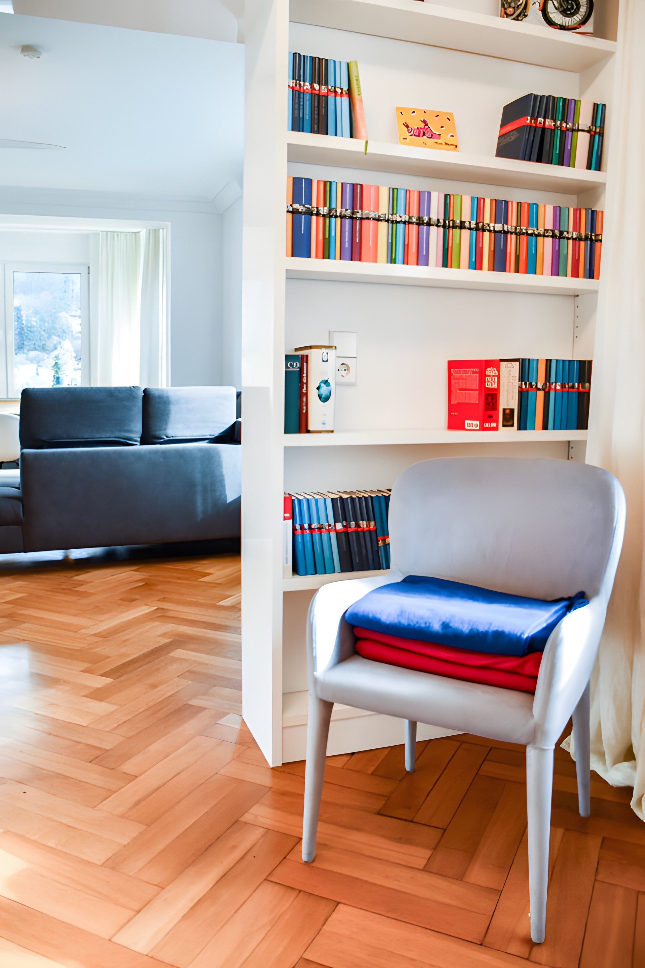 Una sala de estar moderna con un sofá gris, un sillón blanco con un cojín azul y rojo, y una estantería llena de libros y objetos decorativos.