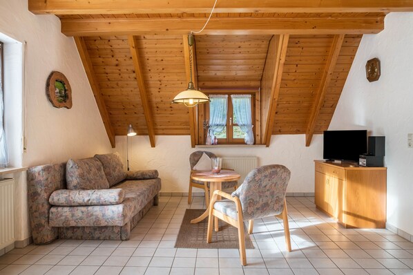 A cozy room with a wooden ceiling, a sofa, a chair, a small table, a television, and a window with curtains.