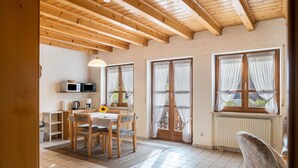 A cozy room with a wooden ceiling, a dining area with a table and chairs, a kitchen with a microwave, and large windows with curtains.