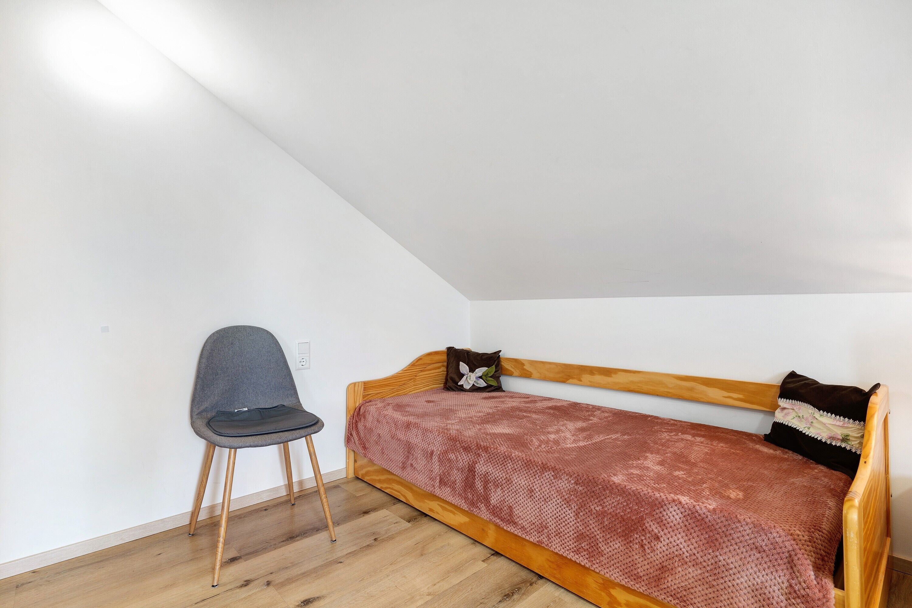 Een kleine eenpersoonskamer met een houten hoofdbord, een grijze stoel en een bed met een decoratief sprei.