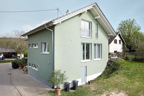 Une maison à deux étages, dotée d’un toit en pente, d’un porche à l’avant et d’un petit jardin devant, agrémenté de plantes en pot.