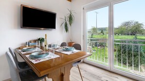 Een eethoek met een houten tafel, stoelen, een aan de muur bevestigde televisie en uitzicht op een tuin door grote ramen.