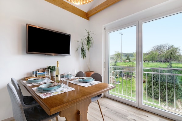 Un coin repas avec une table en bois, des chaises, un téléviseur fixé au mur et une vue sur un jardin grâce à de grandes fenêtres.