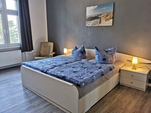 Une chambre à coucher avec un lit, deux tables de chevet, une chaise, une fenêtre avec des rideaux et un tableau au mur.