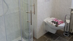 A bathroom with a glass shower enclosure, a toilet with a floral seat cover, and a towel rack. - Apartment "Villa Adler - Waldseite" (Seebad Ahlbeck)