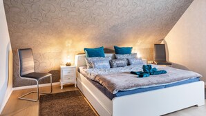 Quarto com teto inclinado, cama com roupa de cama azul e cinza, uma cadeira, mesa de cabeceira e abajur.