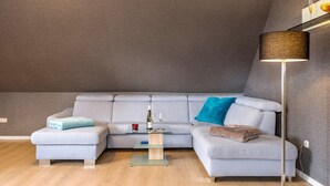 Uma sala de estar moderna com um sofá seccional cinza, uma mesa de centro de vidro, um abajur e uma pequena mesa lateral com uma garrafa e copos.