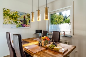 A dining table with a wooden top, surrounded by chairs, with a window view and a painting on the wall.