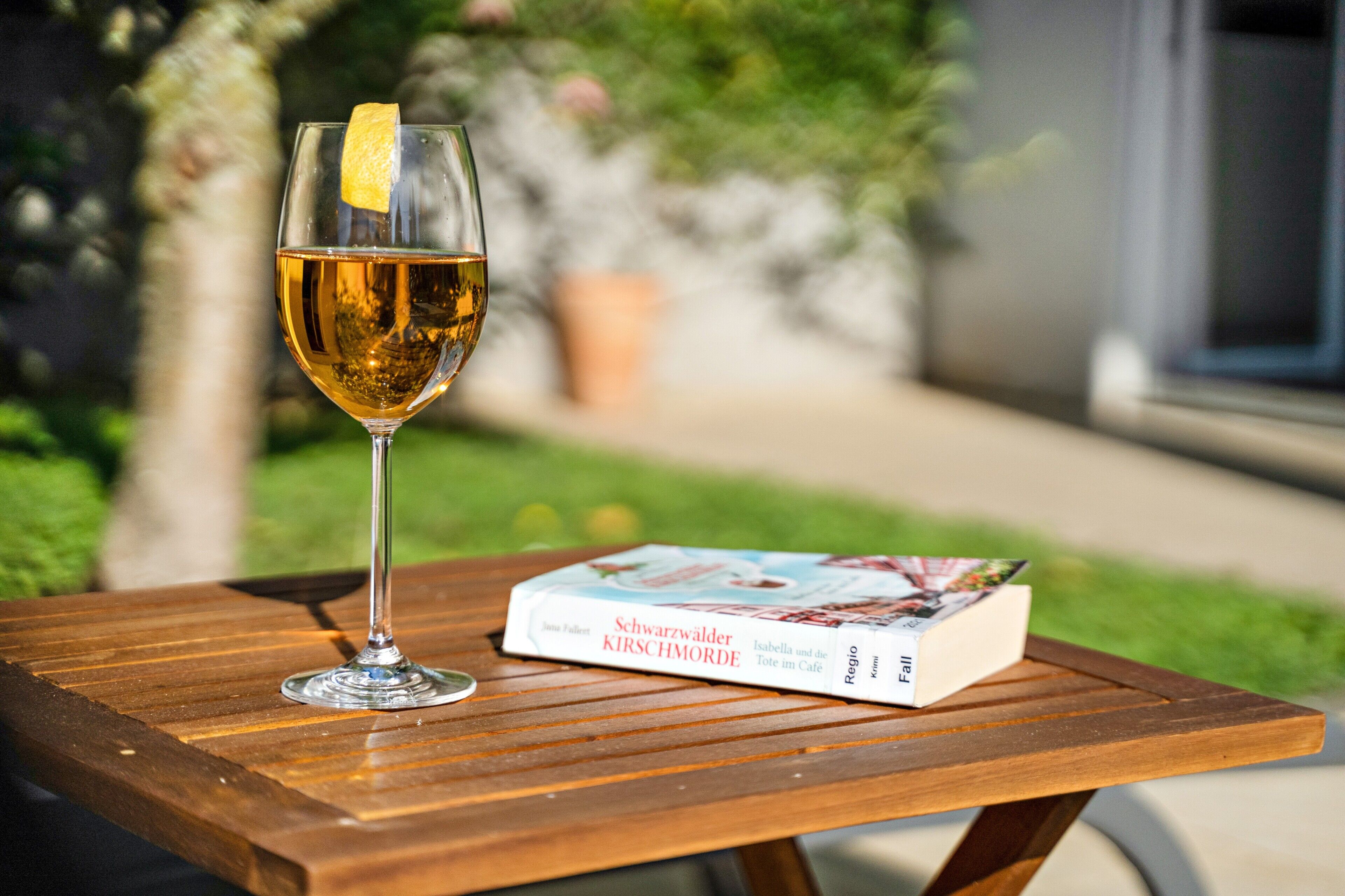 A glass of amber liquid with a lemon peel garnish on a wooden table, next to a book titled 'Schwarzwälder Kirschmorde'.
