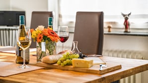 Ein Holztisch mit einer Flasche Weißwein, einem Glas Rotwein, einem Brotlaib, Käse, Trauben und einem Blumenstrauß.