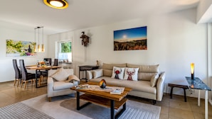 Ein modernes Wohnzimmer mit Sofa, Couchtisch, Esstisch und Stühlen. An der Wand hängt ein Gemälde, auf einem Beistelltisch steht eine Lampe und ein Fenster lässt natürliches Licht herein.