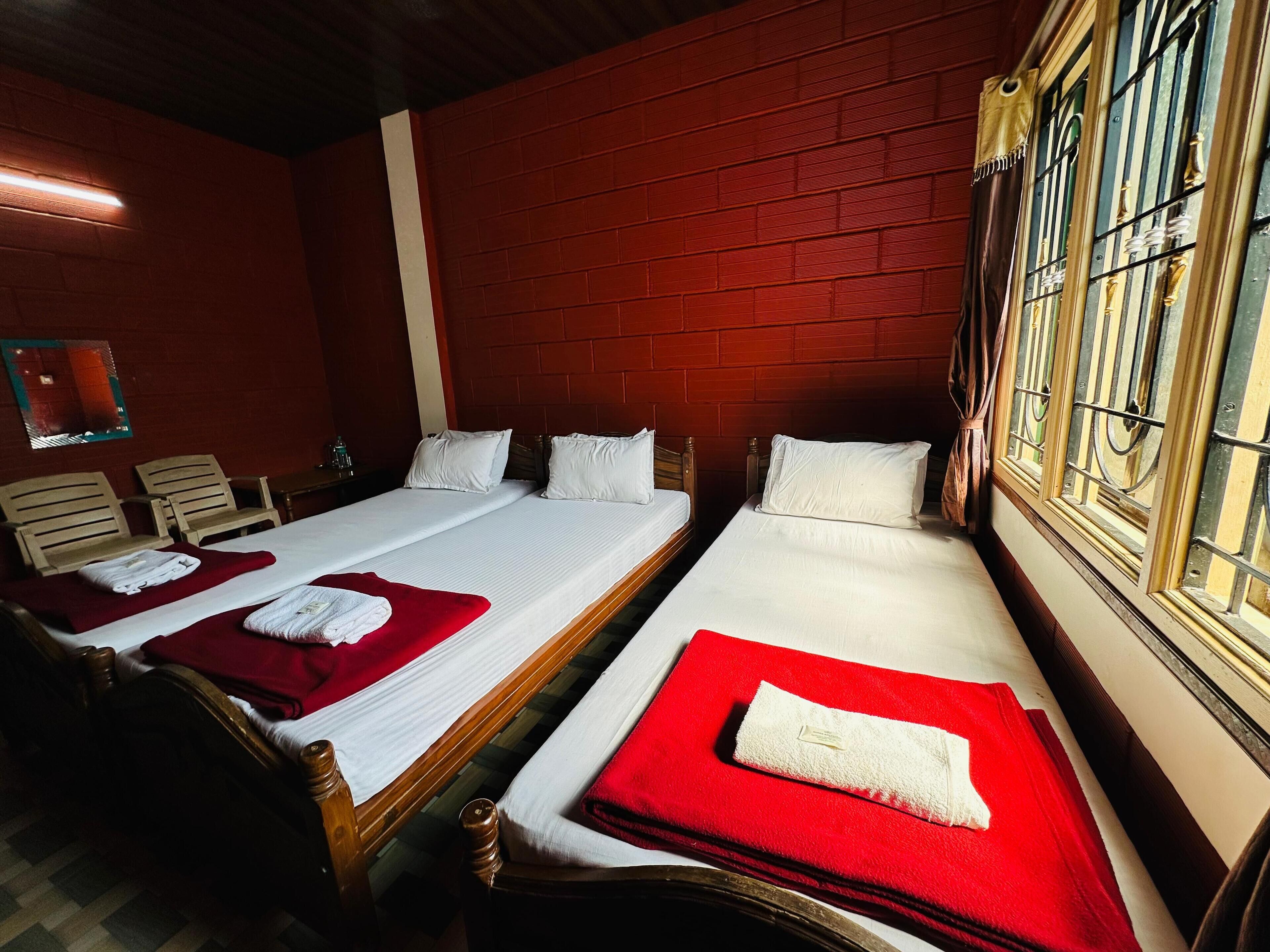A hotel room with two single beds, each with a red blanket and white pillows. The room has a red brick wall and a window with metal bars.