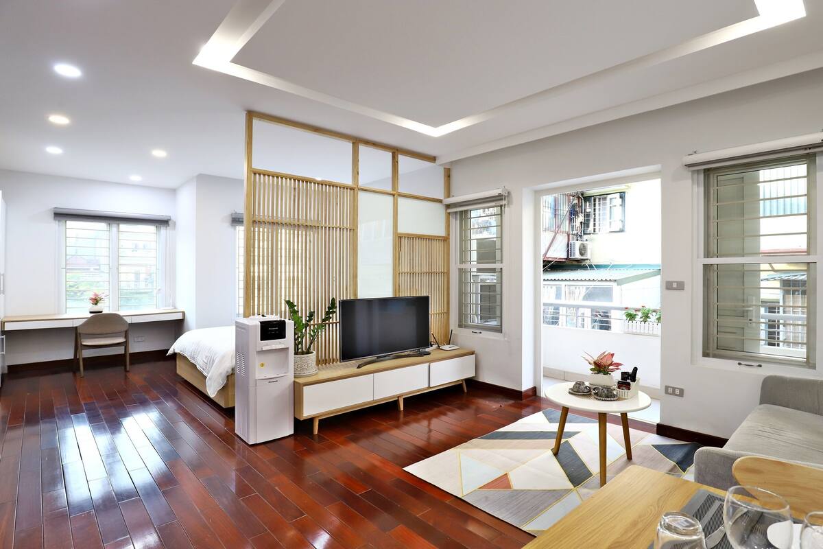 A modern living room with a flat-screen TV, wooden flooring, a sofa, a small round table, and a desk with a chair.