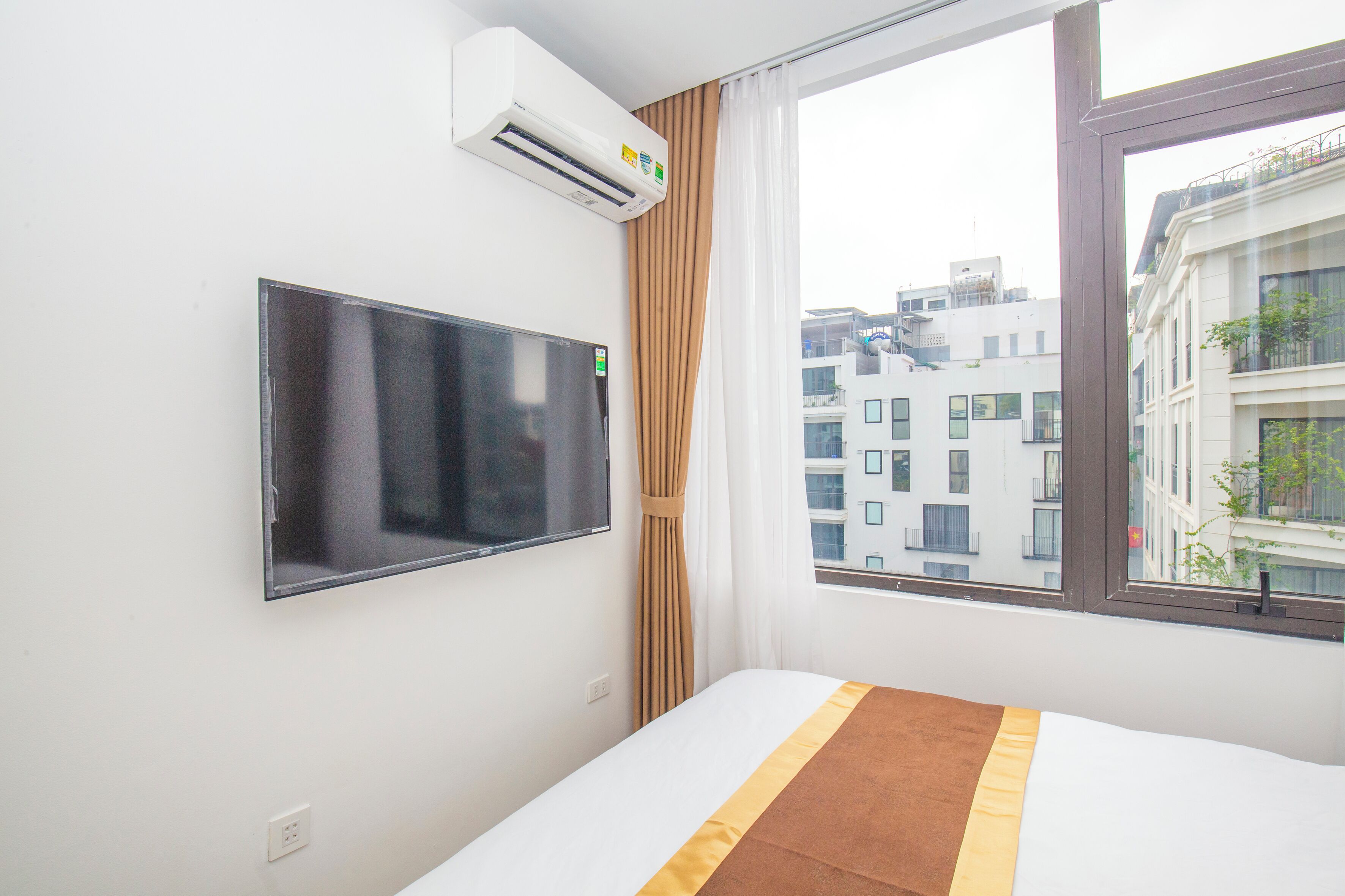 A hotel room with a flat-screen TV mounted on the wall, a window with curtains, and a bed with a brown and yellow striped bedspread.