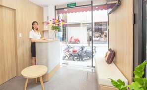 A reception area with a receptionist, a counter, a sofa, a window with a view of a red scooter, and a sign indicating an exit. - Sumitomo13 Apartment   (Hanoi)