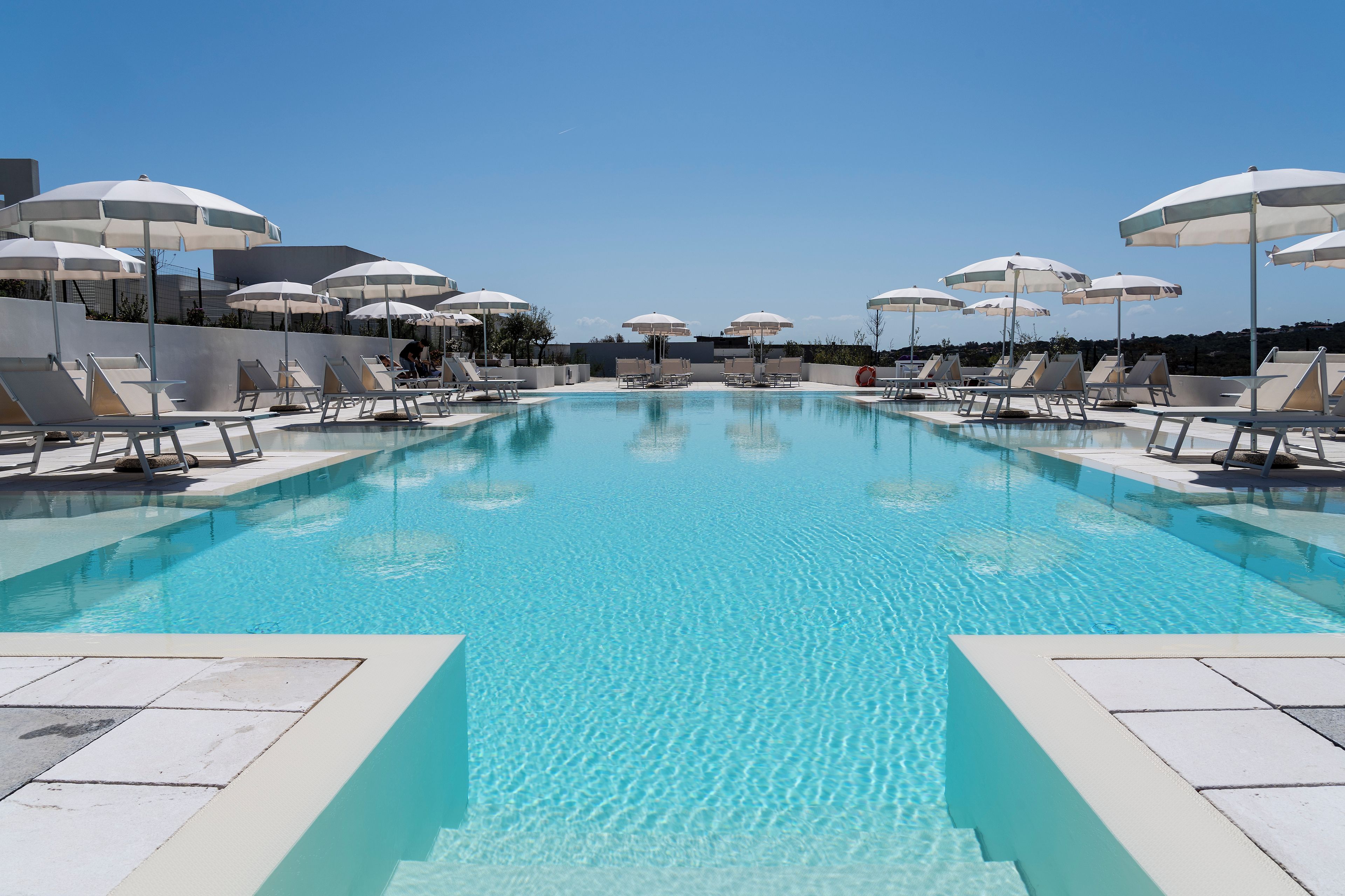 A swimming pool with sun loungers and umbrellas, surrounded by a low wall.