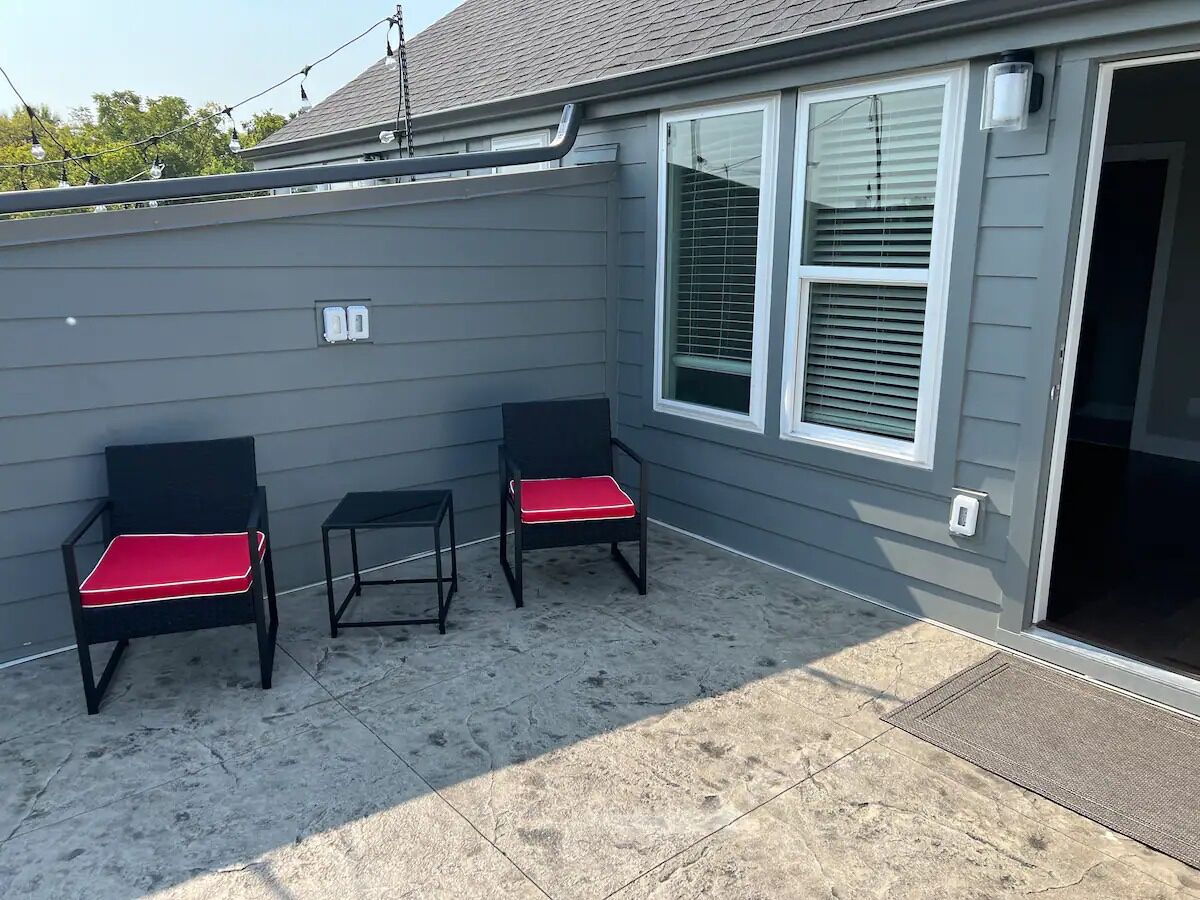 A patio area with two black chairs with red cushions, a small table, and a grey building with a window and door.