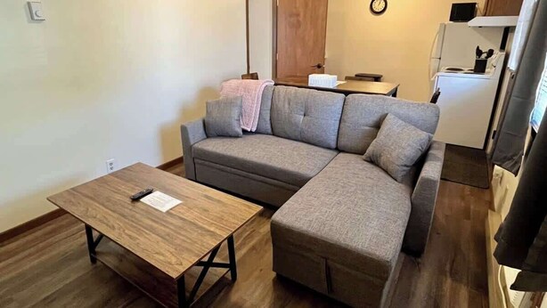 Ett vardagsrum med en soffa, ett soffbord i trä, ett köksutrymme med ett kylskåp och en klocka på väggen.