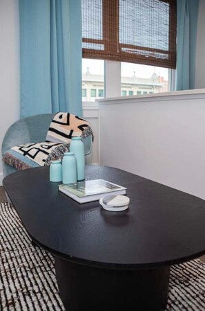 Ein Zimmer mit einem schwarzen runden Tisch, einem blauen Sofa mit gemusterten Kissen und einem Fenster mit Blick auf Gebäude.