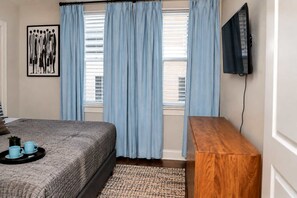 Un dormitorio con cama, una mesita de noche de madera, un televisor montado en la pared y una ventana con cortinas.