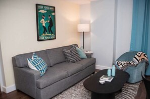 Ein modernes Wohnzimmer mit einem grauen Sofa, gemusterten Kissen, einem schwarzen Couchtisch und einem Poster an der Wand.