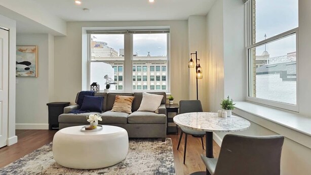 A modern living room with a sofa, ottoman, and round table, large windows with a view of buildings, and a floor lamp.