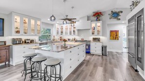 A modern kitchen with a central island, bar stools, a ceiling fan, and a large refrigerator. - Your Hidden Florida Retreat | 4 King Suites, Luxury Kitchen, Heated Pool & Beach Adventures (Fort Myers)
