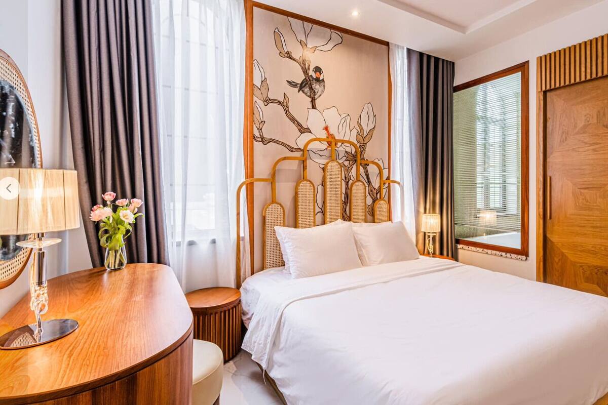 Ein Hotelzimmer mit einem Bett, einem Schreibtisch mit Lampe, einer Vase mit Blumen und einem Wandbild mit Baum und Vogel.