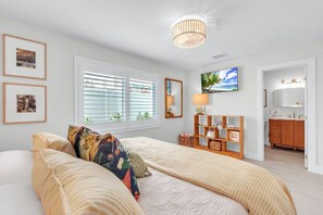 A bedroom with a bed, a bedside table, a mirror, a television, and a window with blinds.