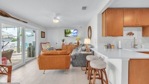 A spacious living area with a sofa, chairs, and a dining area with stools. A kitchen with wooden cabinets and a white countertop is visible.