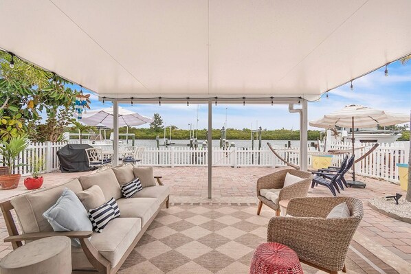 An outdoor patio with a checkered floor, a covered seating area with sofas and chairs, umbrellas, and a view of the water.