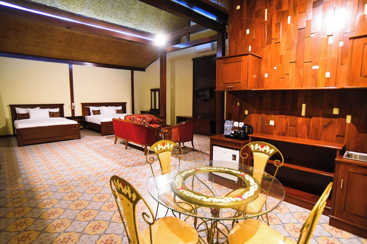 A hotel room with two beds, a red sofa, a glass table with chairs, and wooden paneling on the walls.