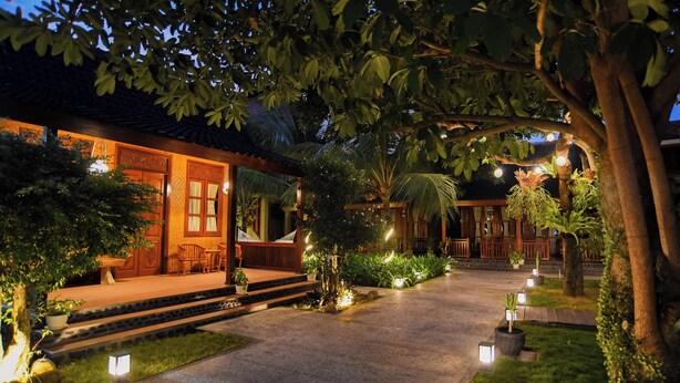 A traditional-style building with a wooden exterior, a covered porch, and a well-lit garden area with potted plants and a pathway. - Ndalem Mataram (Banyuwangi)
