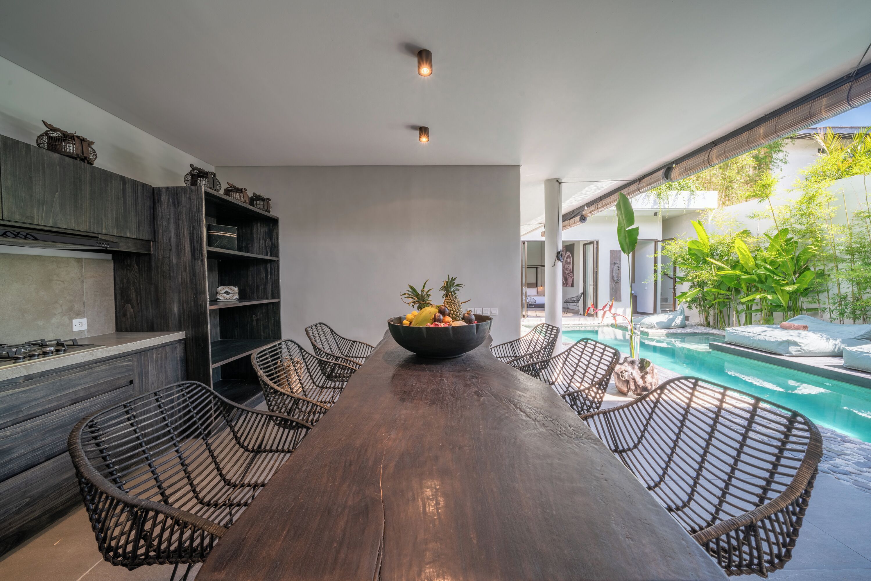 A modern kitchen with dark wood cabinetry, a central island, and a dining area with wicker chairs. A large bowl of fruit is on the island, and a pool is visible in the background.