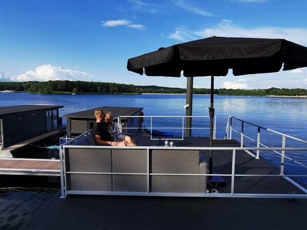 A boat deck with a couple seated under a canopy, a small deck area, and a view of a calm lake with trees in the background.