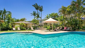 Ein Swimmingpool mit Liegestühlen und Sonnenschirmen, umgeben von Palmen und einem Gebäude mit einer überdachten Galerie.