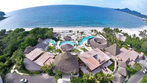Vista aérea de un resort con varias construcciones de techo de paja, una piscina central y una playa al fondo.
