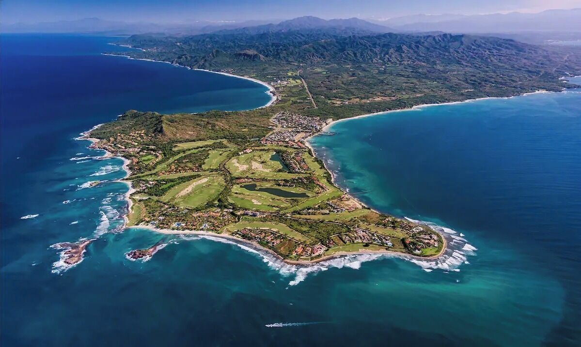 An aerial view of a coastal island with a mix of residential and natural areas, surrounded by clear blue waters and distant mountains.