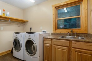 A laundry area with two washing machines, wooden cabinets, a granite countertop, a sink, and a window with a view of trees.