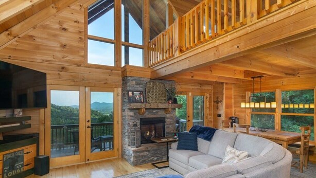 Een gezellig houten chalet interieur met een stenen openhaard, een groot raam met uitzicht op een bos en een houten terras.