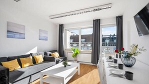 A modern living room with a sofa, a dining table set for two, a large window with curtains, and a flat-screen TV mounted on the wall. - Munich Gem - 2 Bedroom Alpine View Tranquility (München)