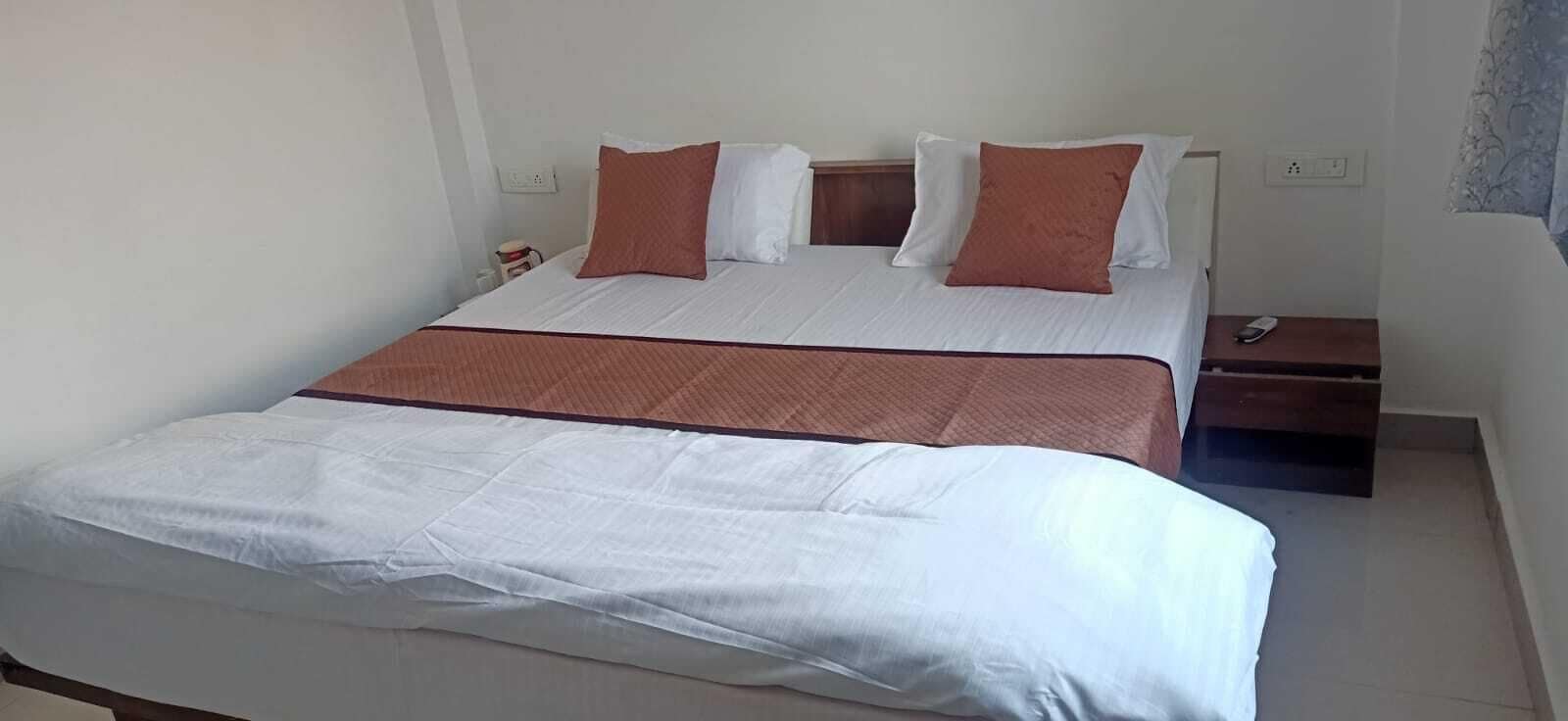 A neatly made bed with white and brown bedding, a bedside table with a remote control, and a power outlet on the wall.