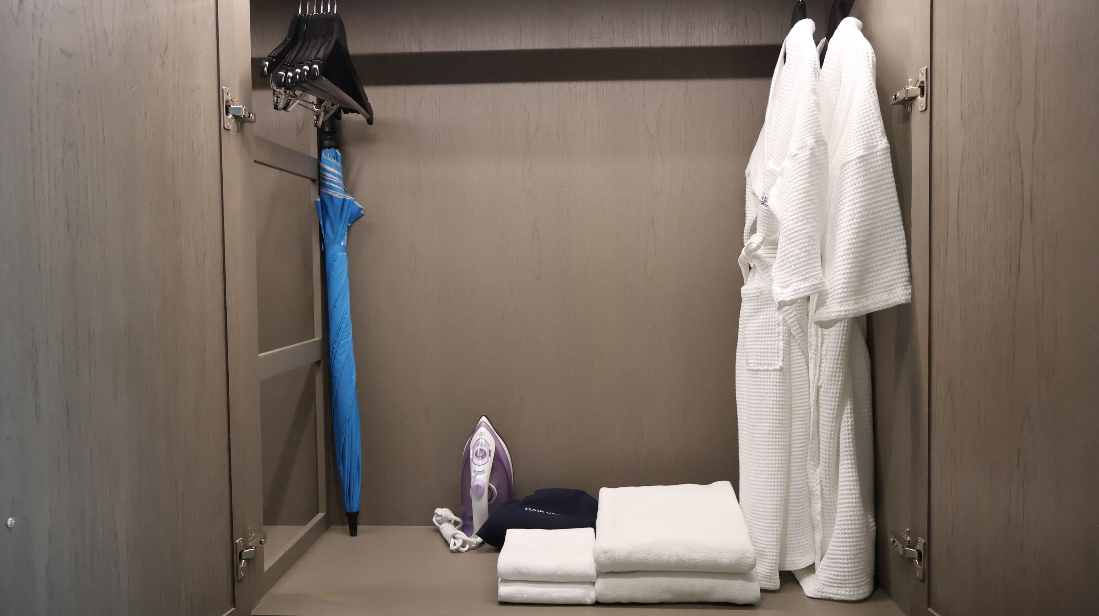 A hotel room with a wardrobe, a blue umbrella, a hairdryer, and a white robe.