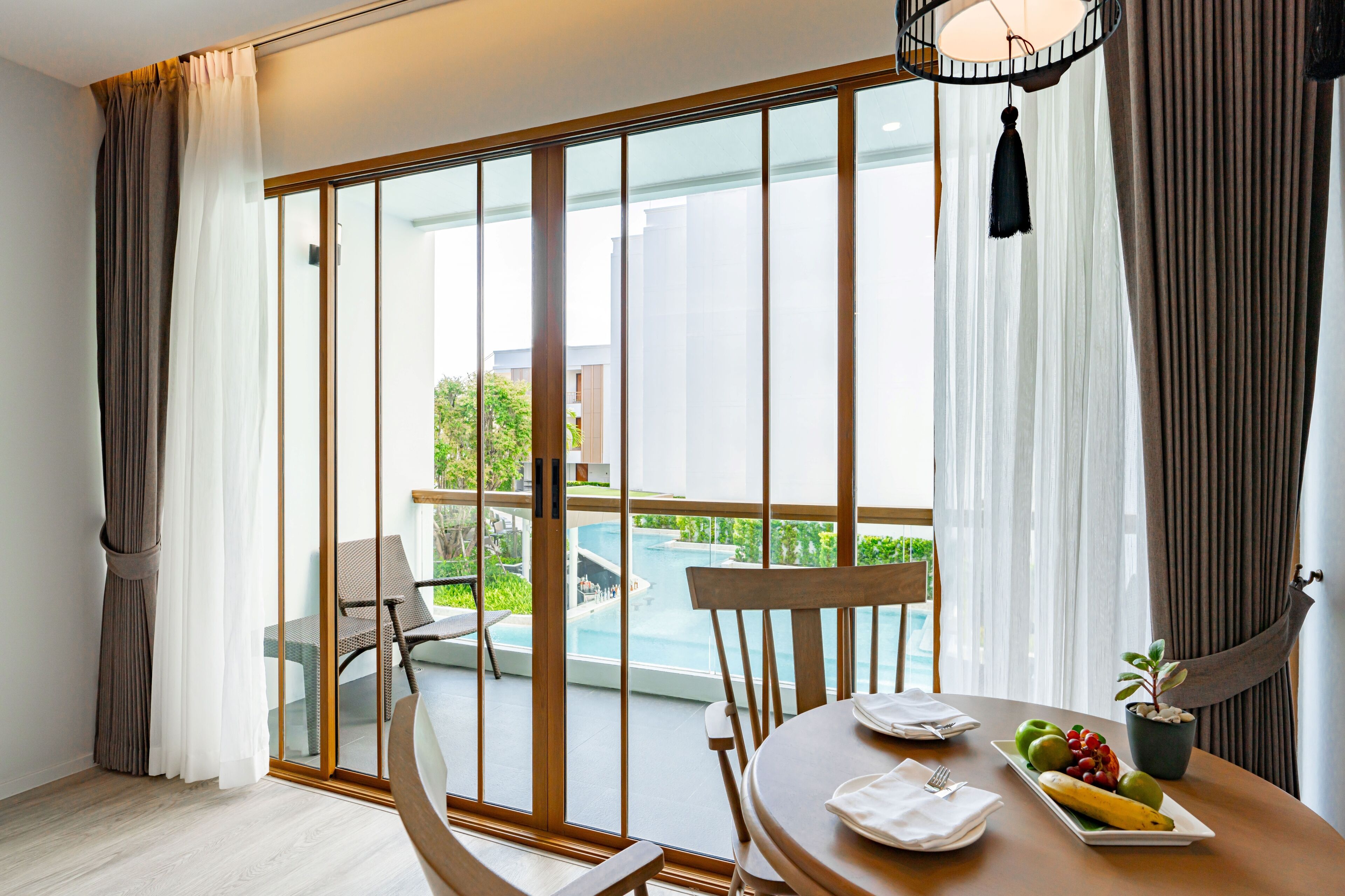 A modern hotel room with a large sliding glass door leading to a pool area. There is a dining table with a fruit plate and a potted plant.