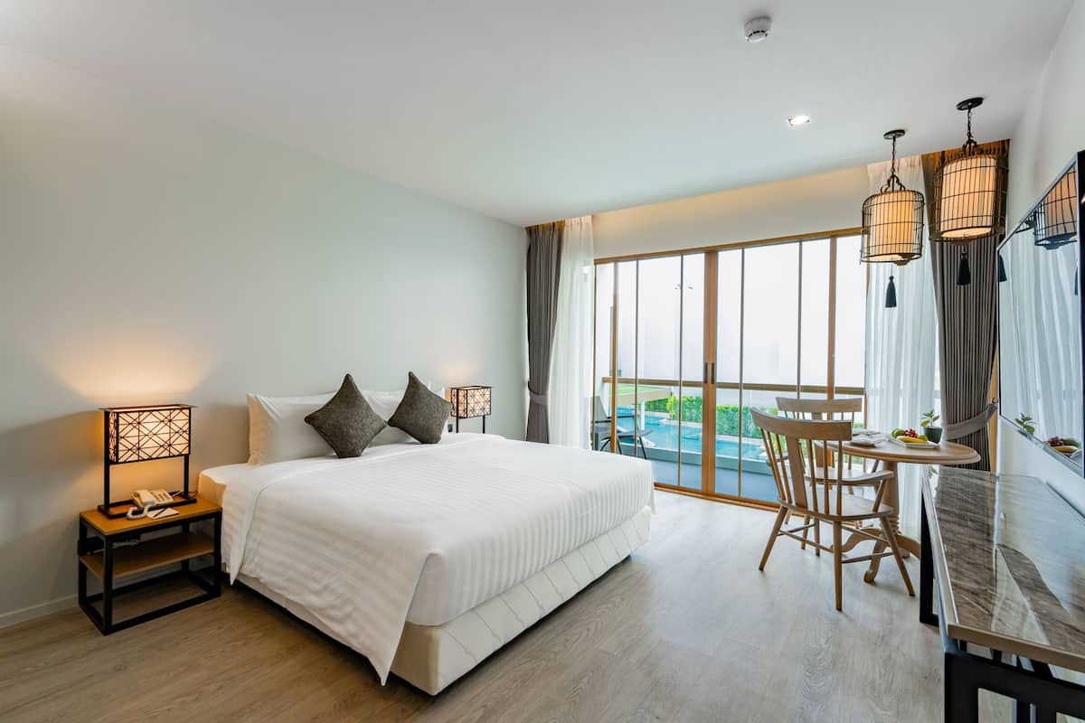 A hotel room with a large bed, bedside table with a lamp, a desk with chairs, and a view of a swimming pool.
