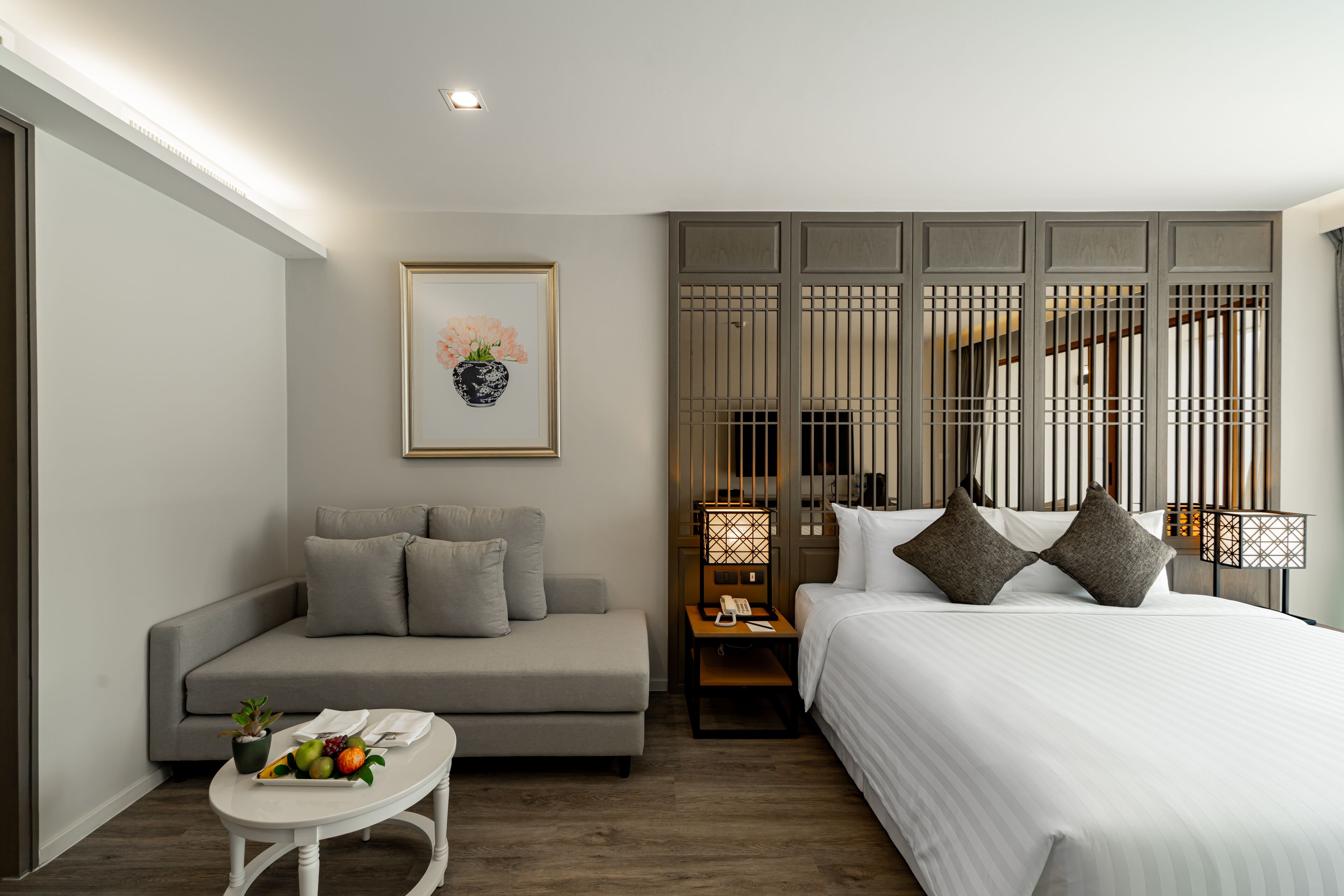 Een moderne hotelkamer met een bank, een klein tafeltje met fruit, een bed met kussens, een nachtkastje met een lamp en een decoratief wandpaneel.
