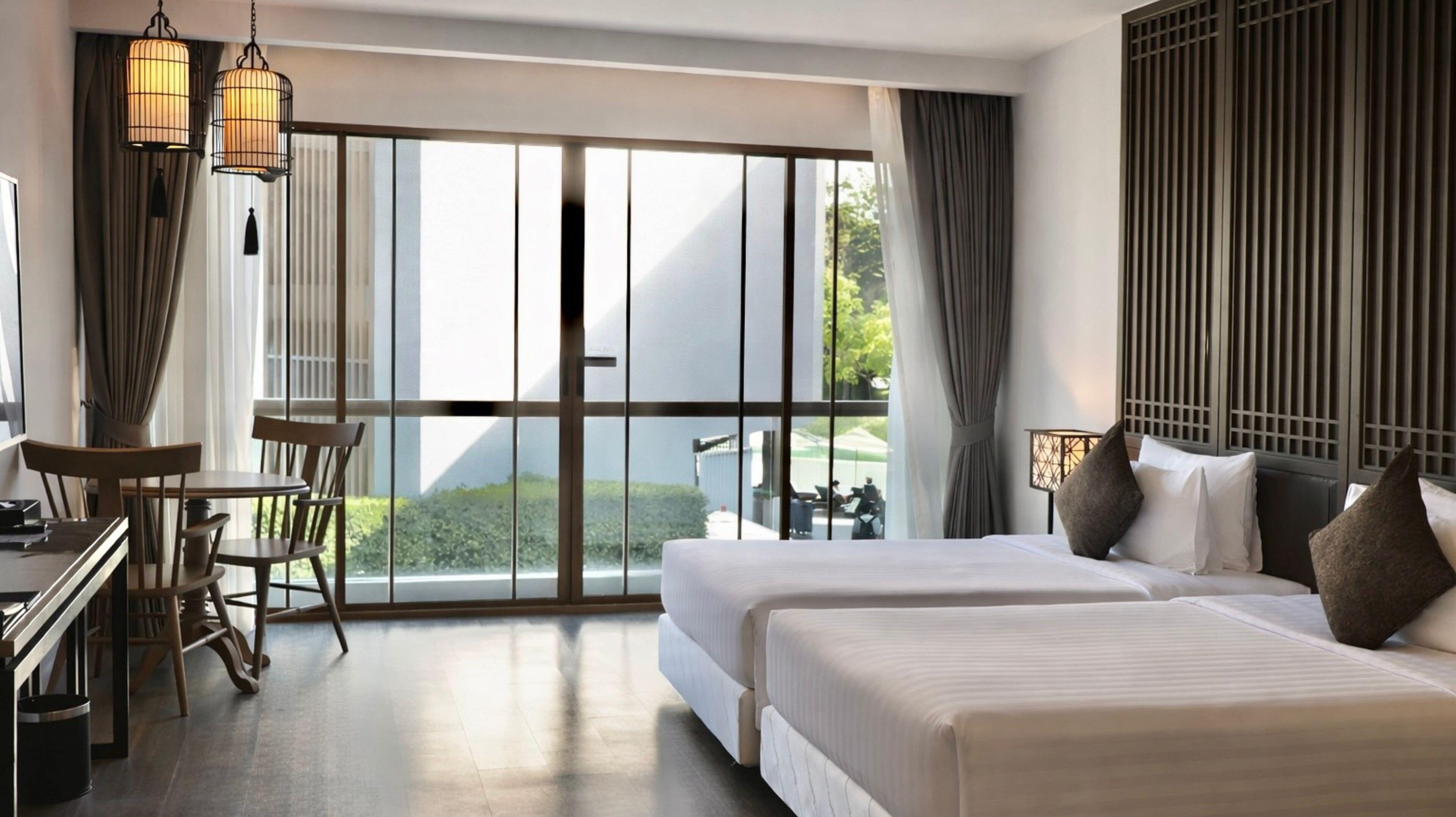 Une chambre d’hôtel dotée d’une grande porte-fenêtre coulissante donnant sur un balcon, d’un lit avec des oreillers, d’un bureau avec une chaise et de deux luminaires suspendus.
