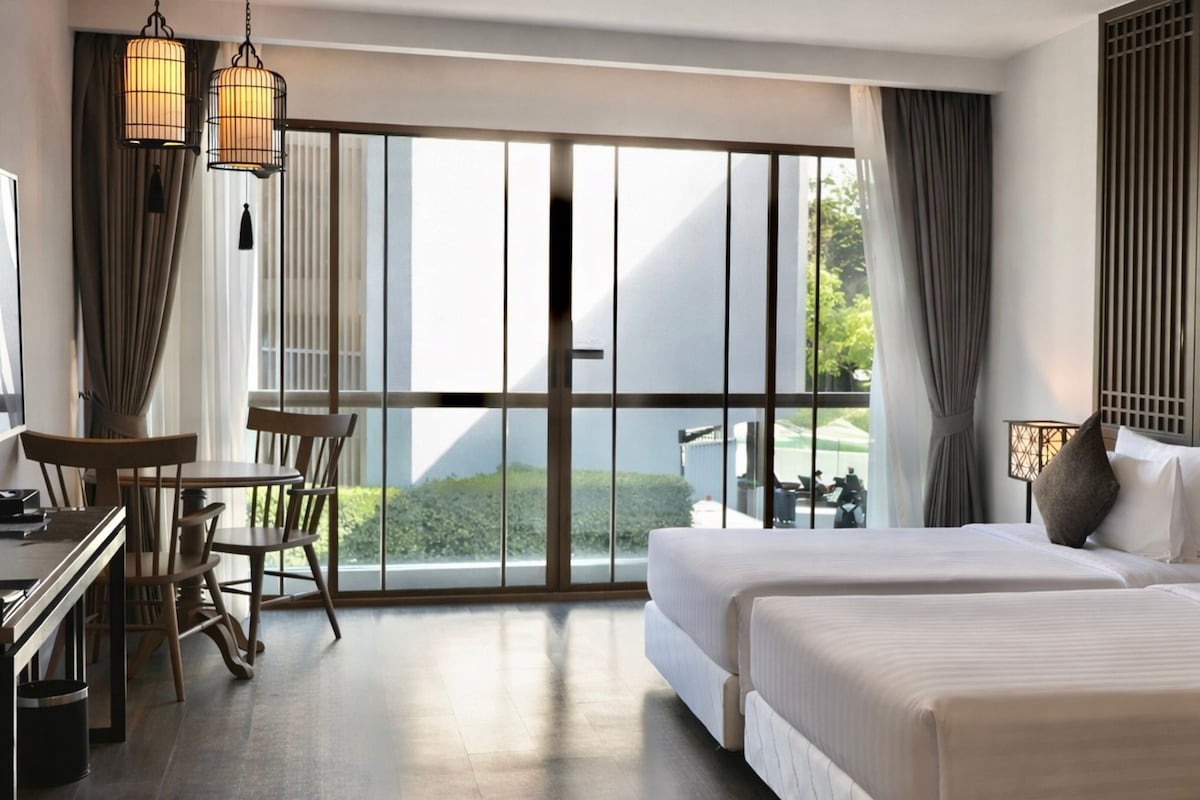 Una habitación de hotel con un gran ventanal corredero que da a un balcón, una cama con almohadas, un escritorio con una silla y dos lámparas colgantes.