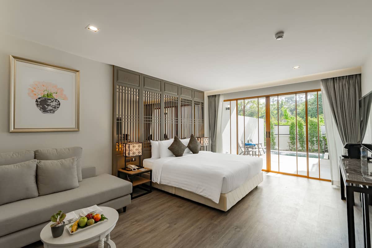 A modern hotel room with a large bed, a sofa, a dining table, and a view of greenery through the sliding glass door.